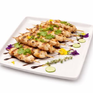 BROCHETTE DE POULET A LA PLANCHA (Minimum de commande 10 brochettes), présentées sur une assiette blanche, garnies de coriandre fraîche, de tranches de concombre, de fleurs comestibles et d'un filet de sauce.