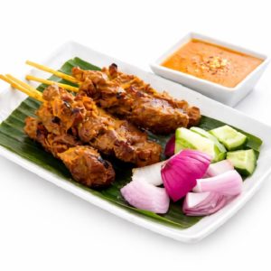 BROCHETTES DE POULET SATAY (Min de commande 10 brochettes) : Servies sur feuille de bananier avec oignons rouges, concombre et sauce orange, le tout présenté sur une assiette rectangulaire blanche.