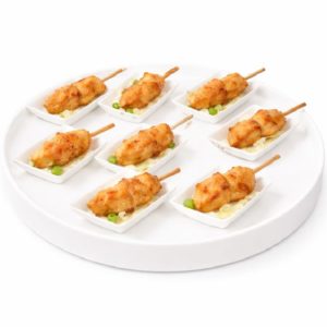 Huit brochettes de BROCHETTE POULET PIC & CHIC, panées et de la taille d'une bouchée, chacune servie avec une sauce jaune et une garniture verte dans de petits plats blancs, disposés sur un plateau rond blanc.
