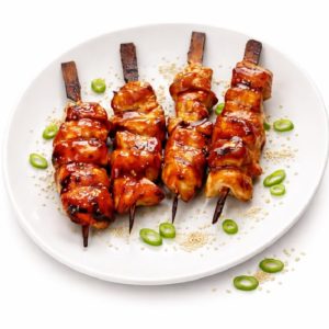 Quatre brochettes de BROCHETTE POULET PIC & CHIC, glacées et grillées, servies sur une assiette blanche avec des graines de sésame et une garniture d'oignons verts. Commande minimum : 10 brochettes.