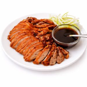 CANARD LAQUÉ DE PEKIN (250 g/pers) servi sur une assiette blanche avec des tranches de canard croustillant, un bol en verre de sauce foncée avec une cuillère, et une garniture d'oignons verts finement tranchés.