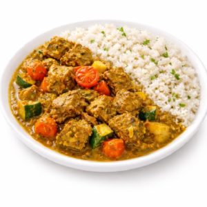 Assiette blanche avec du riz blanc garni d'herbes, accompagné de COLOMBO DE PORC - curry de porc avec courgettes, carottes, pommes de terre et tomates cerises dans une sauce épaisse. Commande minimum : 5 pièces (300g chacune).