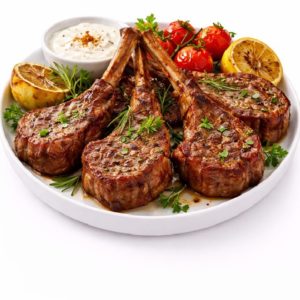 CÔTE D'AGNEAU CUITE AU FEU DE BOIS (Barquette de 5 pièces), servies grillées avec herbes, tomates cerises rôties, citrons et sauce crémeuse sur assiette blanche.