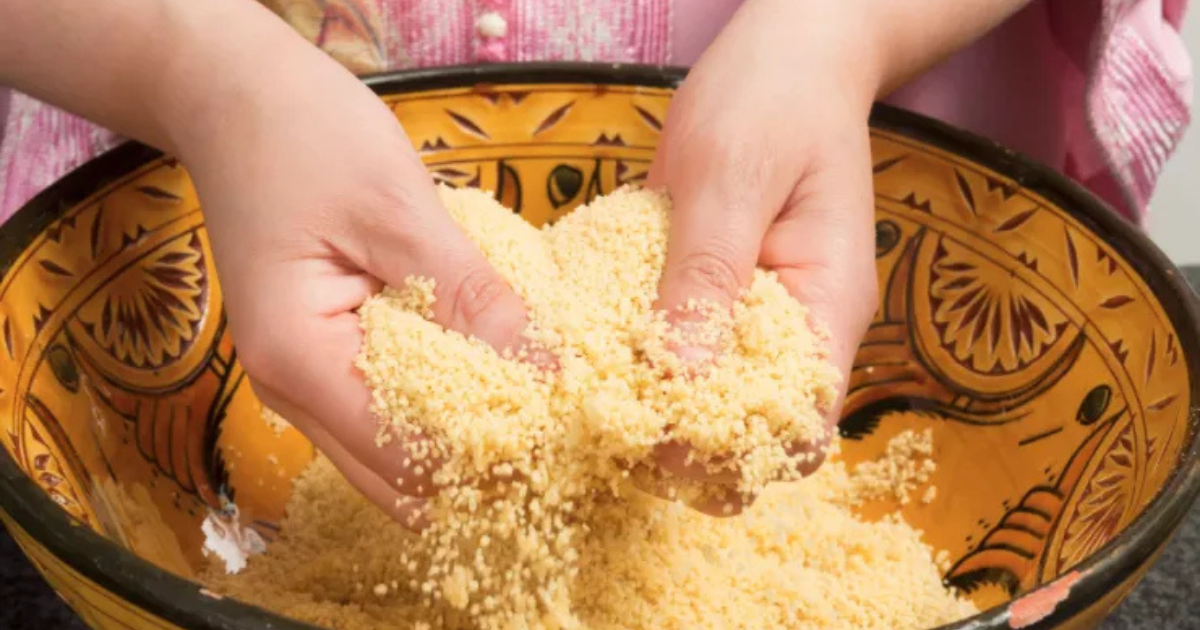 Une personne utilisant ses deux mains pour mélanger ou ébouriffer du couscous sec dans un bol décoratif à motifs jaunes et bruns.