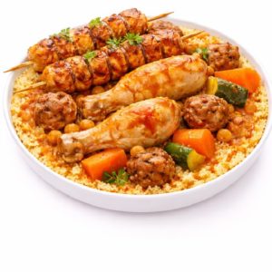 Une assiette blanche avec COUSCOUS BROCHETTES DE POULET, servi avec couscous, pilons de poulet, boulettes de viande, pois chiches, carottes, courgettes et garniture d'herbes fraîches.
