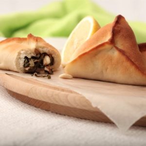 Trois FATAYER ÉPINARDS (80 g chacun), dont l'un est ouvert pour révéler une garniture d'épinards et de pignons, reposent sur une planche en bois parcheminée, avec un quartier de citron en arrière-plan. Commande minimum : 5 pièces. Entrée chaude.