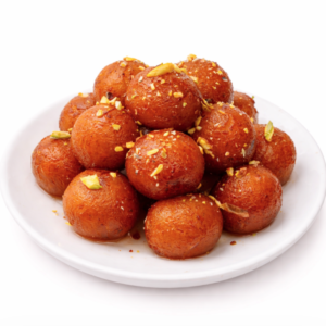 Une assiette blanche avec GULAB JAMUN (Barquette de 10 pièces) - des gâteaux ronds et dorés garnis de pistaches hachées - sur un fond blanc uni.