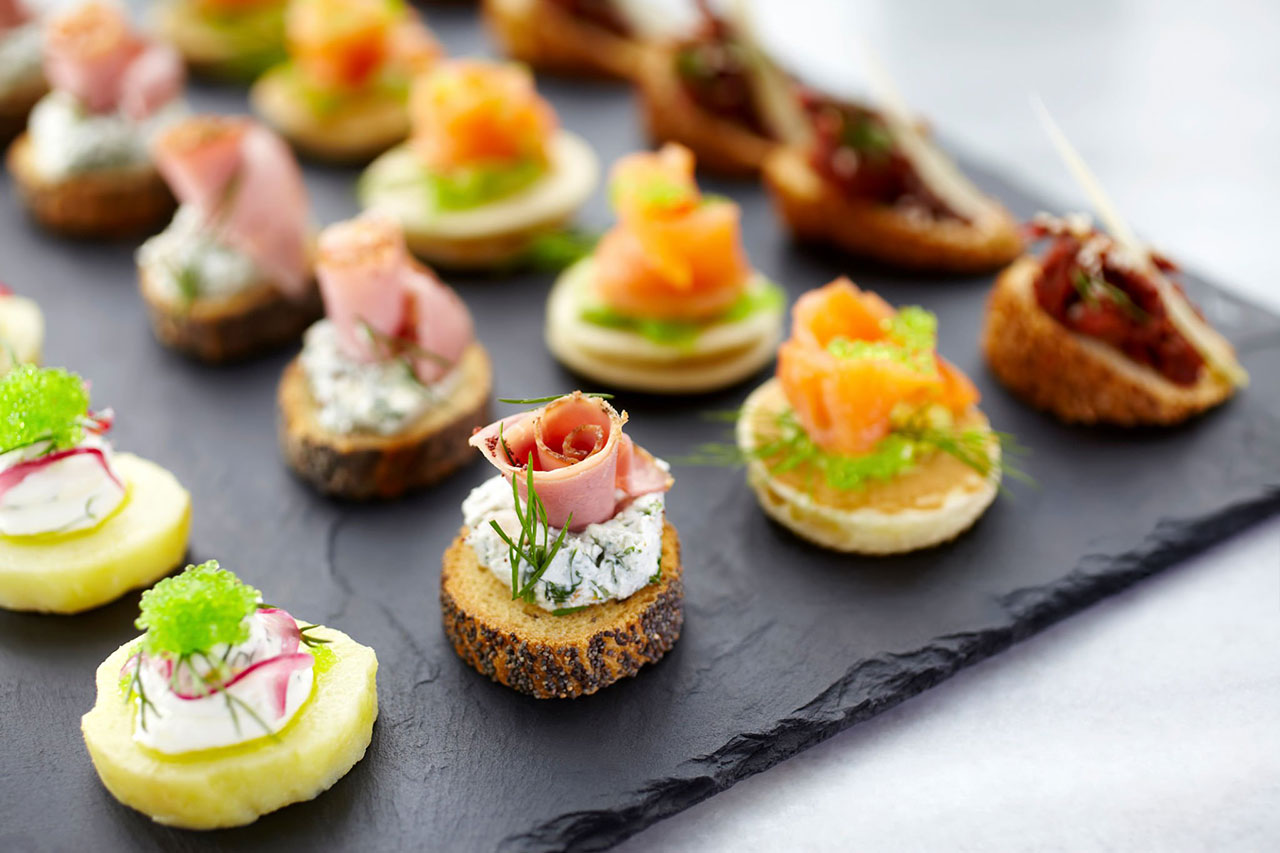 Un assortiment de canapés gastronomiques colorés, y compris des tranches de pain garnies de jambon, de saumon, d'herbes et de crème, disposés proprement sur un plat de service en ardoise noire.
