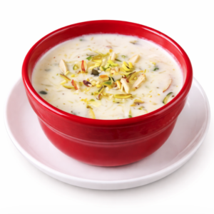 Un bol de KHEER (La part), riz au lait crémeux indien garni d'éclats de pistaches et d'amandes, servi dans une assiette blanche sur fond blanc.