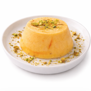 Une portion de KULFI MANGO (La pièce), un dessert glacé traditionnel indien, est présentée sur une assiette blanche sur fond blanc, garnie de pistaches hachées.