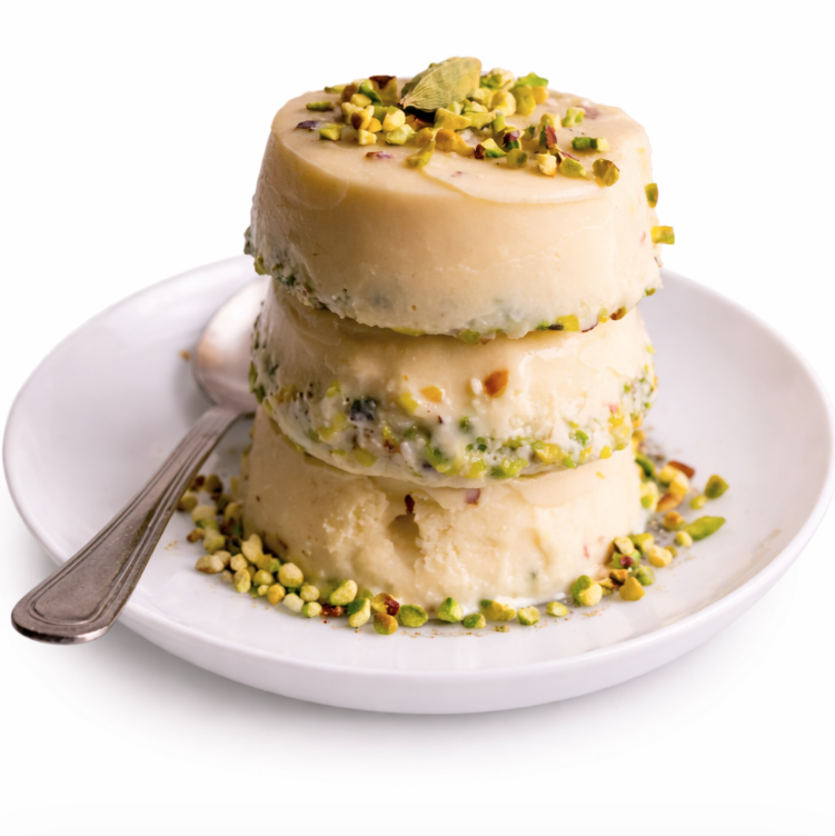 Trois morceaux ronds de PISTACHE KULFI (La pièce) sont empilés sur une assiette blanche, garnis de pistaches hachées et d'une gousse de cardamome, avec une cuillère à côté.