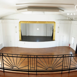 Salle d'événements spacieuse et vide avec des planchers en bois, une petite scène encadrée d'or, de grandes fenêtres et une balustrade noire de style art déco. L'inscription "LE LOFT SOLARIS (75020)" figure dans le coin supérieur droit.