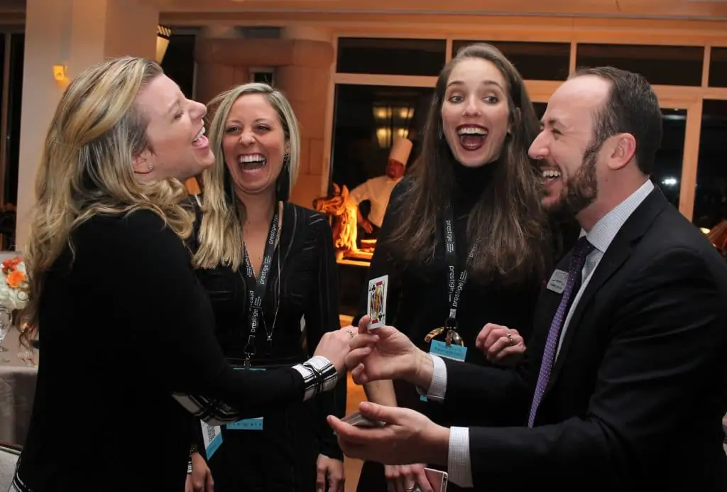 Un groupe de trois femmes et un homme rient ensemble à l'intérieur tandis que l'homme tient une carte à jouer. Ils semblent être à un événement formel ou à une fête, et tout le monde a l'air surpris et joyeux.