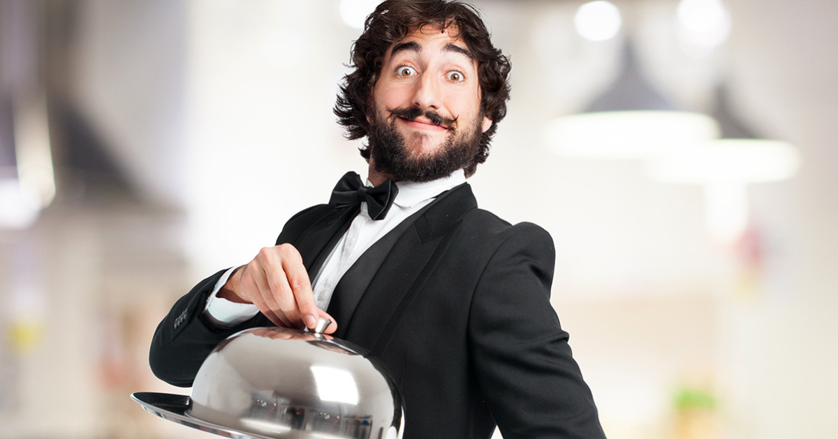 Un serveur souriant en smoking noir tenant un plateau de service en argent avec un couvercle bombé, debout dans un restaurant très éclairé.