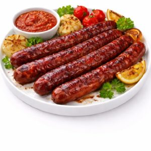 Dix saucisses MERGUEZ CUITE AU FEU DE BOIS servies sur une assiette blanche avec de l'ail rôti, des tomates cerises, des quartiers de citron, du persil et un bol de sauce rouge.
