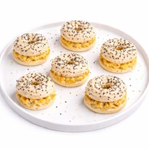 Dix MINI BAGELS POULET CURRY (20 g chacun, commande minimum) sont disposés sur une assiette ronde blanche. Les mini-bagels sont fourrés au poulet au curry et recouverts de graines de pavot et de sésame. Quelques miettes sont éparpillées sur l'assiette.
