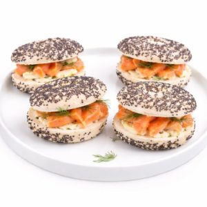 Les MINI BAGELS SAUMON (20g/pièce, minimum 10) sont fourrés de saumon fumé, de fromage frais et d'aneth frais, servis dans une assiette ronde blanche et garnis d'aneth supplémentaire.