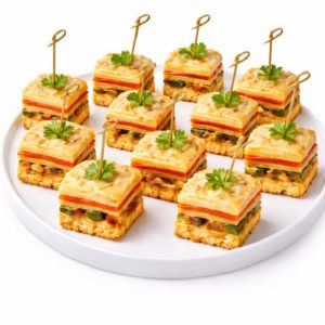 Une assiette blanche présente dix MINI LASAGNES LÉGUMES - des amuse-gueules carrés à base de légumes et de pain, chacun garni de persil et maintenu par un pic décoratif.