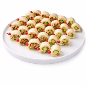 Une assiette ronde blanche contient le PLATEAU DE 48 MINI WRAPS, des rouleaux de tortilla de la taille d'une bouchée, soigneusement disposés et remplis de légumes et d'autres ingrédients, chacun étant fixé par un cure-dent à pointe rouge sur un fond blanc.
