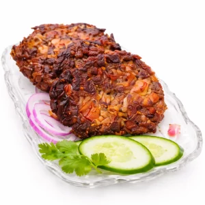 Le NARGISHI SHEEK KABAB (300 g) présente deux galettes brunes croustillantes sur une assiette en verre, garnies d'oignons rouges émincés, de concombre et de coriandre, sur fond blanc.