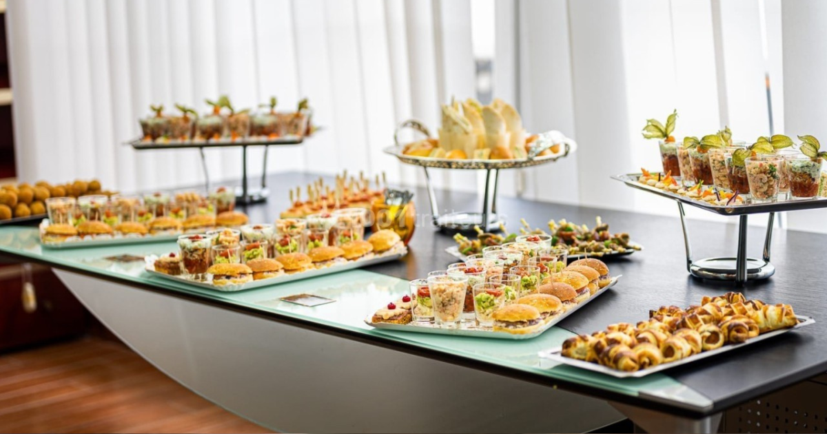 Une table de buffet avec un assortiment d'amuse-gueule, y compris des sandwichs, des pâtisseries, des brochettes et des salades dans de petites coupes, disposés proprement sur des plateaux et des supports dans une pièce lumineuse avec des stores verticaux.