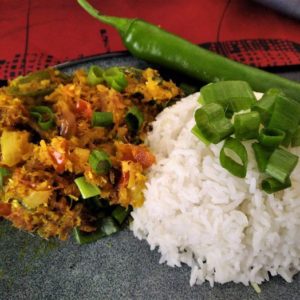 Une assiette de ROUGAIL DE MORUE (commande minimum de 5 plats, 300g/personne) servie avec du riz blanc, des oignons verts hachés, du curry de légumes et un piment vert sur le côté sur une nappe rouge et noire.