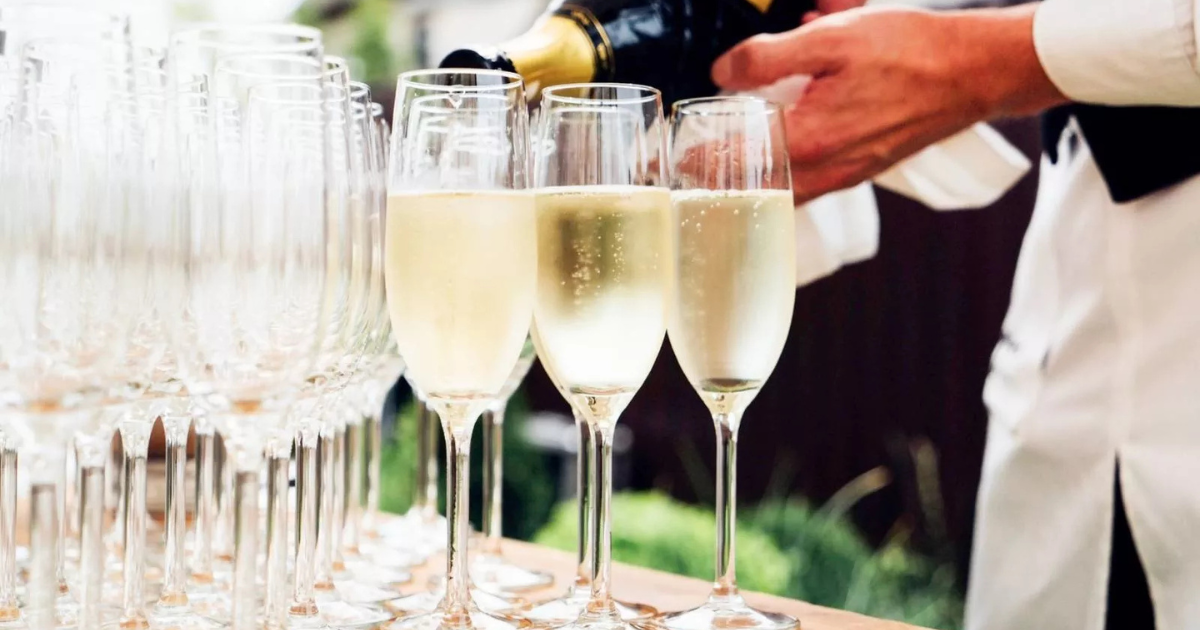 Une personne verse du vin mousseux ou du champagne dans plusieurs grands verres disposés en rangées sur une table, avec d'autres verres vides à l'arrière-plan.