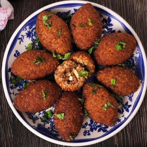 Une assiette de huit KEBBÉ (Entrée chaude - Min de commande 5 pièces - 80g/pièce), croustillants et de forme ovale, garnis d'herbes hachées. L'un d'eux est ouvert, montrant sa viande épicée et sa garniture de pignons de pin sur une surface en bois sombre.