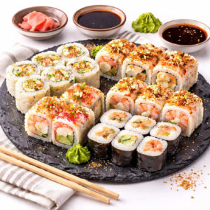 Le PLATEAU SUSHIS TOUT CUIT est servi sur un plateau rond noir avec un assortiment de rouleaux de sushi, garnis d'assaisonnements colorés. La sauce soja, le wasabi et le gingembre mariné se trouvent dans de petits bols à proximité, avec des baguettes sur une serviette rayée.