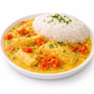 Une portion de VIVANEAU À LA CRÈME DE CURCUMA (commande minimum de 5 plats, 250 g chacun) comprend du riz blanc et une sauce crémeuse au curcuma jaune avec du poulet, des tomates cerises et des herbes fraîches sur une assiette blanche.