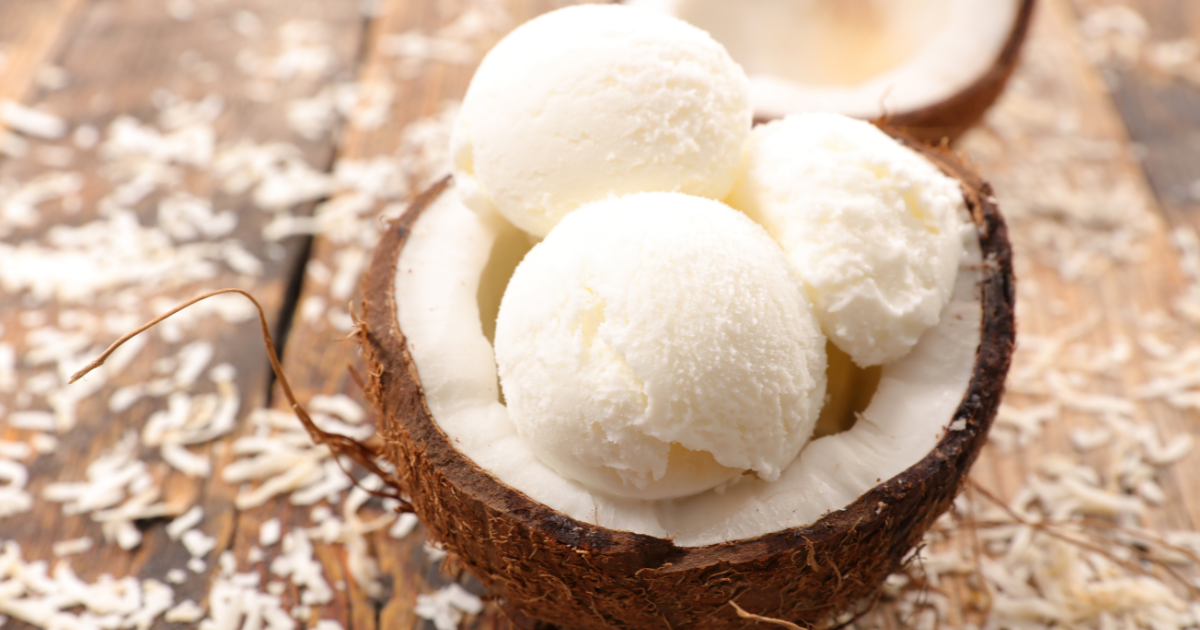 Trois boules de glace à la vanille sont servies dans une coquille de noix de coco coupée en deux, entourée de noix de coco râpée éparpillée sur une surface en bois rustique.