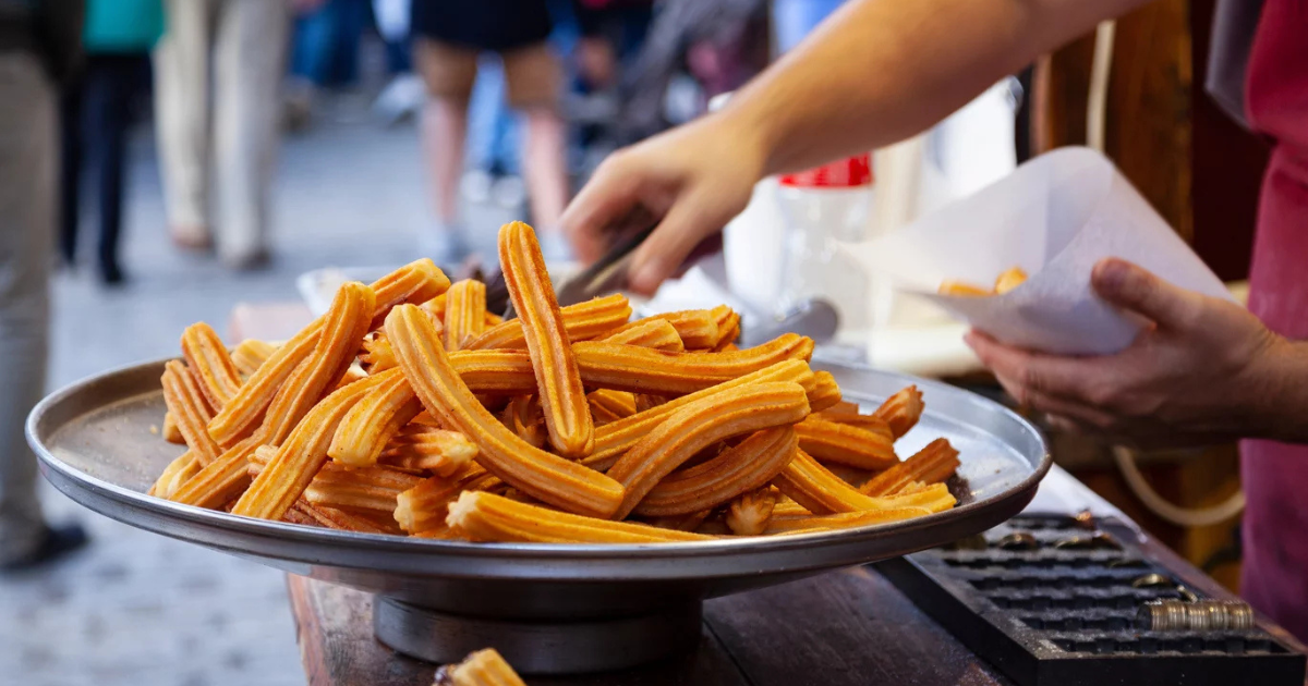 Un grand plateau rempli de churros frais est posé sur un comptoir en bois. Une personne utilise des pinces pour prendre des churros et les placer dans un cornet en papier, avec une foule floue en arrière-plan.