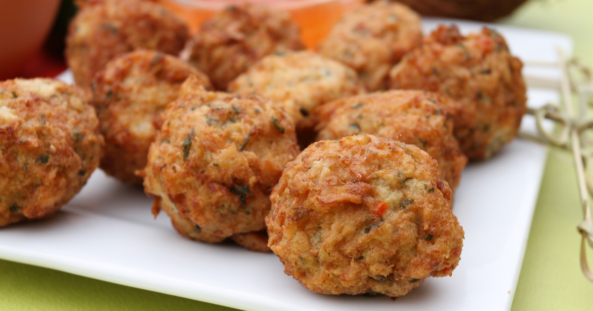 Gros plan sur plusieurs boulettes de viande frites et dorées, parsemées d'herbes, disposées sur une assiette blanche. L'arrière-plan est légèrement flou, avec des touches d'orange et de vert.