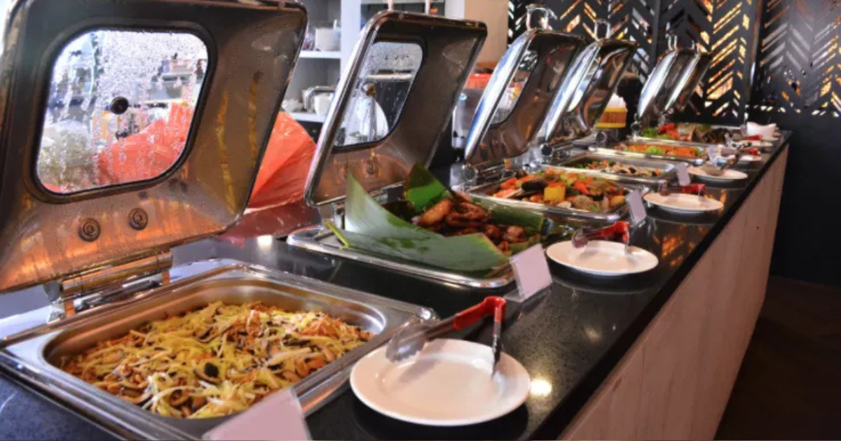 Un buffet composé de plusieurs chafing dishes contenant une variété d'aliments chauds, accompagnés d'ustensiles de service et d'assiettes vides disposés devant chaque plat.