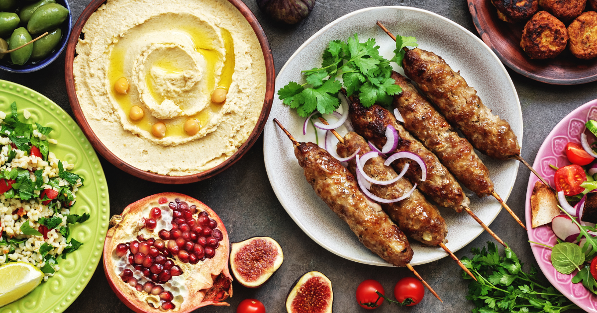 Une assiette colorée de produits du Moyen-Orient, comprenant du houmous à base de pois chiches et d'huile d'olive, des brochettes de viande grillée avec des oignons et des herbes, de la salade, des falafels, des figues, de la grenade, des olives et des tomates cerises.