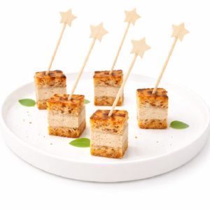Cinq petits canapés TRESOR CUBE FOIE GRAS sont servis sur une assiette blanche, chacun étant embroché sur un bâtonnet étoilé et garni de feuilles vertes.
